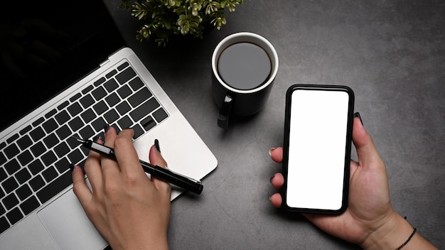 Desde arriba, una mujer que usa una computadora portátil y sostiene un teléfono móvil con una pantalla vacía