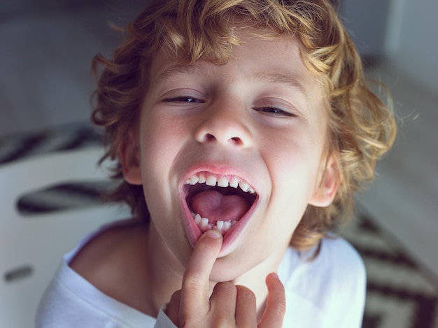 Desde arriba de un niño alegre con cabello rubio ondulado mostrando la boca abierta con un diente de leche perdido mientras toca el labio y mira la cámara