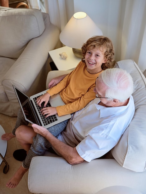 Desde arriba de un niño alegre con su abuelo escribiendo en un netbook mirando a la cámara con una sonrisa mientras está sentado en un sillón en casa
