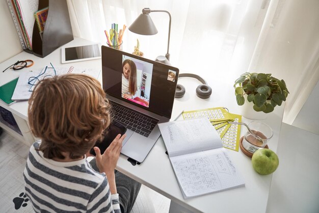 Foto desde arriba, un niño chatea por video con un compañero de clase en una computadora portátil en la mesa con un libro de ejercicios mientras estudian juntos en la habitación