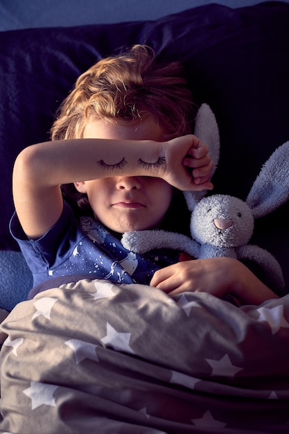 Desde arriba de un niño cubriéndose la cara con la mano con los ojos cerrados pintados mientras finge dormir en la cama con un conejito de peluche