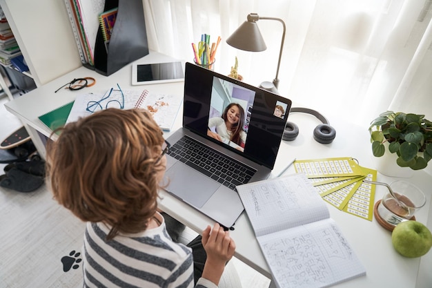 Desde arriba, un niño diligente haciendo video chat con un compañero de clase en una computadora portátil en la mesa con un libro de ejercicios mientras hacían la tarea juntos