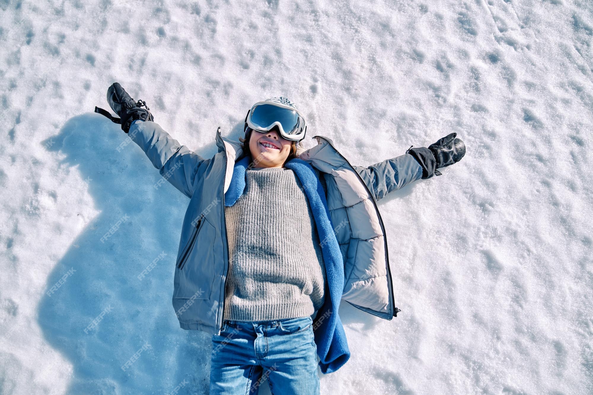 Desde arriba de un niño feliz con ropa abrigada y gafas de esquí  descansando sobre la nieve blanca con los brazos levantados