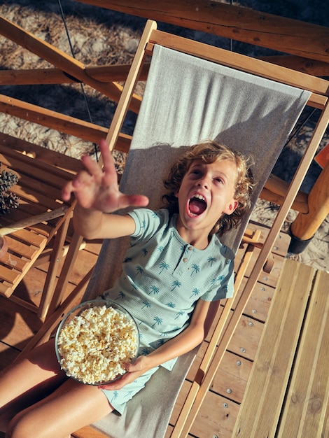 Foto desde arriba de un niño positivo con un tazón de palomitas de maíz gritando con el brazo levantado mientras está sentado en una tumbona en la terraza del campo