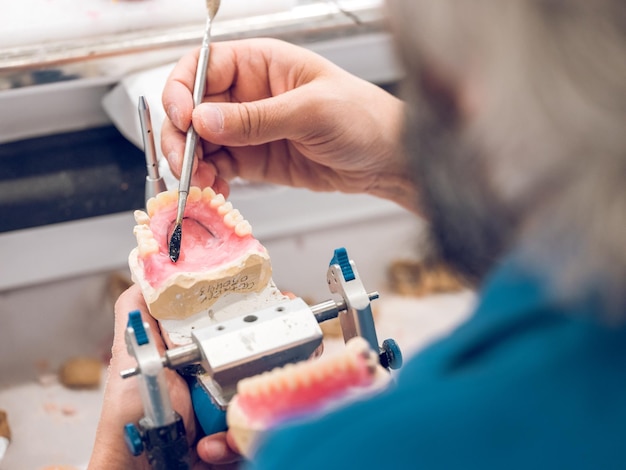 Foto desde arriba del ortodoncista masculino sin rostro usando una espátula dental para procesar y dar forma a la dentadura postiza mientras trabaja en el laboratorio