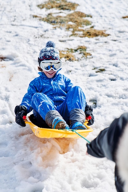 Desde arriba, un padre tirando de un trineo de plástico amarillo con un niño alegre vestido de abrigo mientras se divierte en una pendiente nevada en invierno