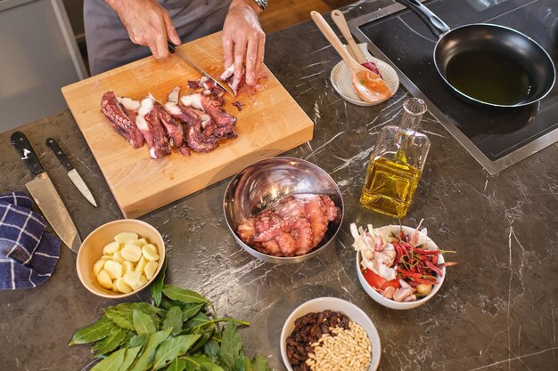 Desde arriba de una persona anónima cortando pulpo crudo en una tabla de madera mientras cocina en la cocina