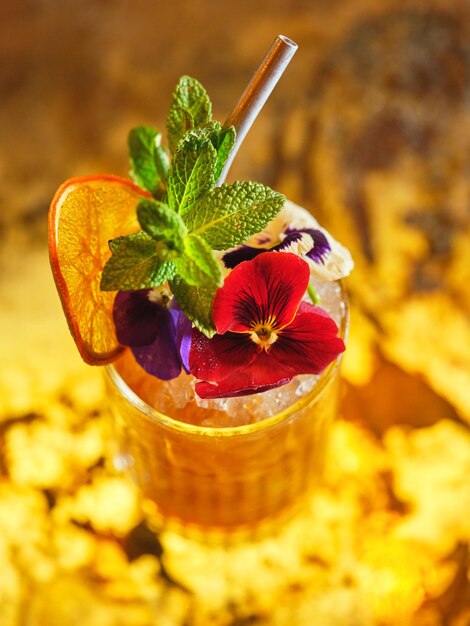 De arriba vaso de bebida alcohólica decorado con flor comestible y rebanada de naranja seca servido con hojas de menta en la barra