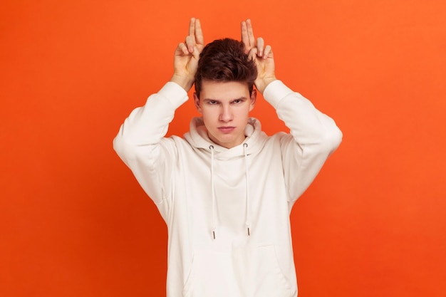 Foto arroganter wütender teenager in modischem weißem hoodie, der hörner zeigt, die die finger in der nähe des kopfes halten, hartnäckiger mann, der charaktermissbraucher demonstriert studioaufnahme isoliert auf orangefarbenem hintergrund