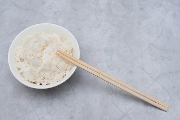 Arroz blanco cocido en tazón blanco con palitos de madera sobre fondo gris Enfoque selectivo Concepto de comida asiática