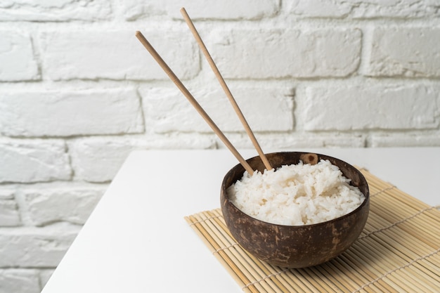 Arroz branco em uma tigela de coco em um fundo branco. foto minimalista com pauzinhos de arroz e bambu.