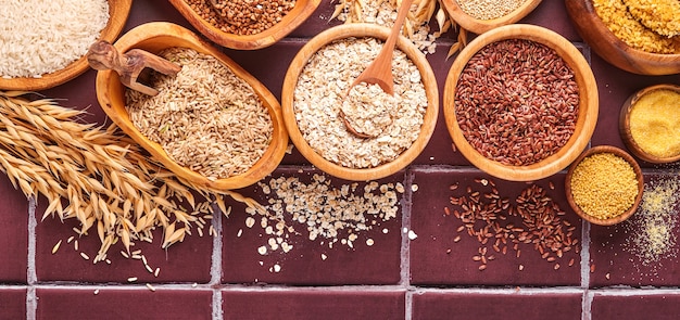 Arroz branco, marrom e vermelho, trigo sarraceno, painço, sêmola de milho, quinua e bulgur em tigelas de madeira na mesa cinza claro da cozinha. Cereais sem glúten. Vista superior com copyspace. Bandeira.