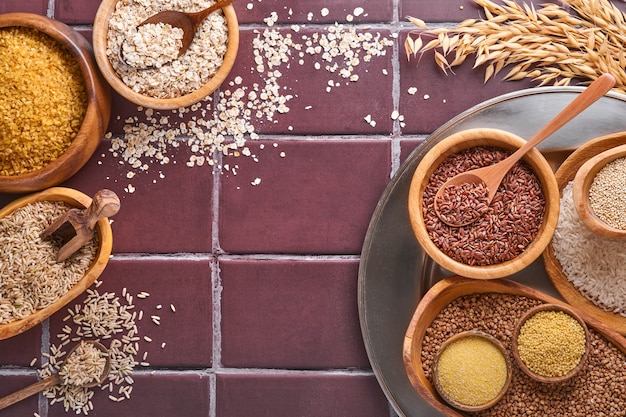 Arroz branco, marrom e vermelho, trigo sarraceno, painço, sêmolas de milho, quinua e bulgur em tigelas de madeira sobre uma mesa de cozinha de pedra marrom. Cereais sem glúten. Vista superior com copyspace