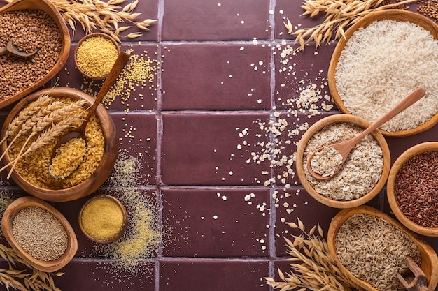 Arroz branco, marrom e vermelho, trigo sarraceno, painço, sêmolas de milho, quinua e bulgur em tigelas de madeira sobre uma mesa de cozinha de pedra marrom. Cereais sem glúten. Vista superior com copyspace