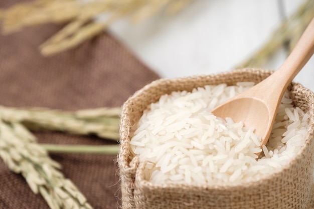 Foto arroz y cuchara de madera en saco con planta de arroz amarillo sobre fondo de madera.