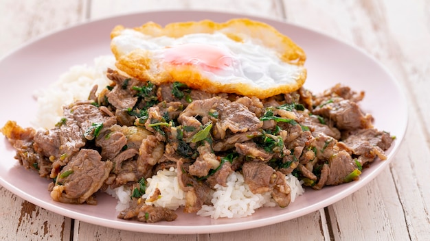 Arroz fluído com carne frita com folhas de manjericão, pimenta longa, pimenta longa indiana, pimenta longa javanesa, pimenta, alho, pimenta e ovo frito em prato de cerâmica com fundo de textura de madeira branca