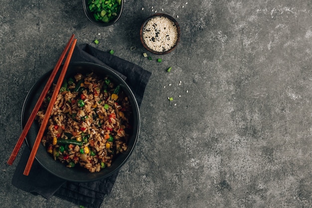 Arroz frito com legumes, cebola verde e gergelim em uma tigela preta sobre fundo de pedra.