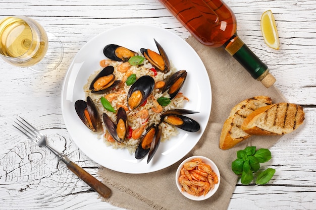 Arroz frito com mexilhões de frutos do mar, camarões e manjericão em um prato com um copo de vinho, serapilheira e baguete torrada na mesa de madeira rachada branca. Vista do topo.