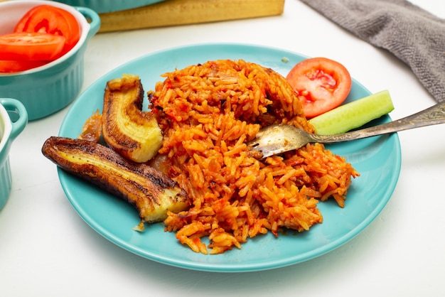 Arroz jollof con plátano frito en un plato Arroz con tomates cebollas especias Verduras frescas Fondo blanco