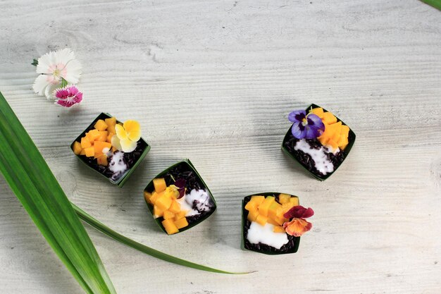 Arroz pegajoso caseiro de coco preto com manga no copo de folhas pandan, foco seletivo com cobertura de flores de edibel amores-perfeitos, espaço para cópia no topo