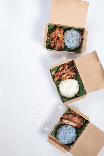Foto arroz pegajoso com porco grelhado e porco frito colocado em uma caixa de papel pardo, coloque sobre uma toalha de mesa branca, caixa de comida, comida tailandesa.