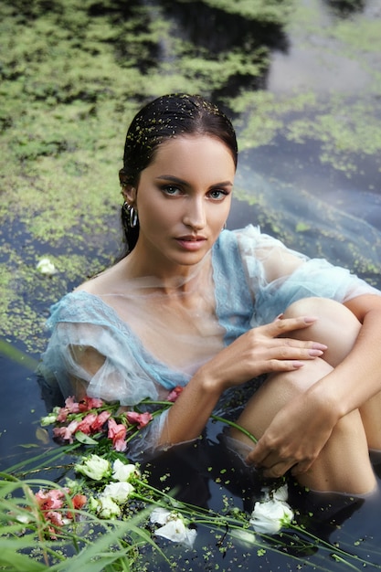 Arte bela mulher romântica encontra-se no pântano em um vestido longo azul com flores. Morena de retrato em vestido transparente em lentilha d'água de lama de pântano. Capa de livro