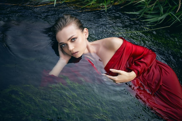 Arte belo retrato romântico de uma jovem sexy em um vestido vermelho deitado em um rio com algas verdes no verão na natureza durante o dia