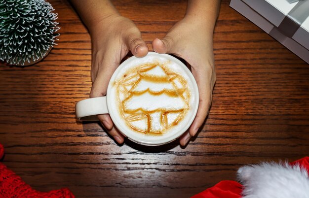 Foto arte do café com leite em forma de árvore de natal com xarope castanho em xícara de café com a mão segurando ao redor