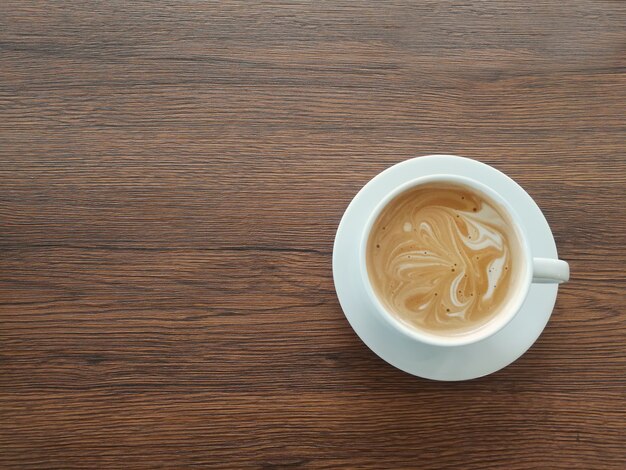 El arte de espuma de café mocha es de mármol en una taza blanca, colocada a la derecha una mesa de madera marrón.