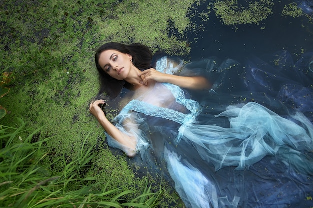 Arte hermosa mujer romántica se encuentra en pantano en vestido largo azul con flores. Morena de retrato en vestido transparente en lenteja de agua de barro de pantano de agua. Tapa del libro