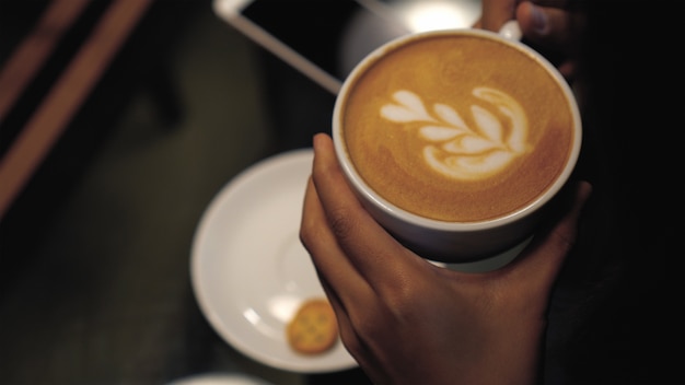 Arte del latte del primer en tapa en tazas de café de la mano de la mujer que se sostiene en la cafetería