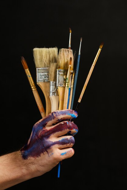 Foto arte moderna e criatividade. ferramentas e suprimentos de pintura. o close up do bando de pincéis na mão masculina sobre o fundo escuro.