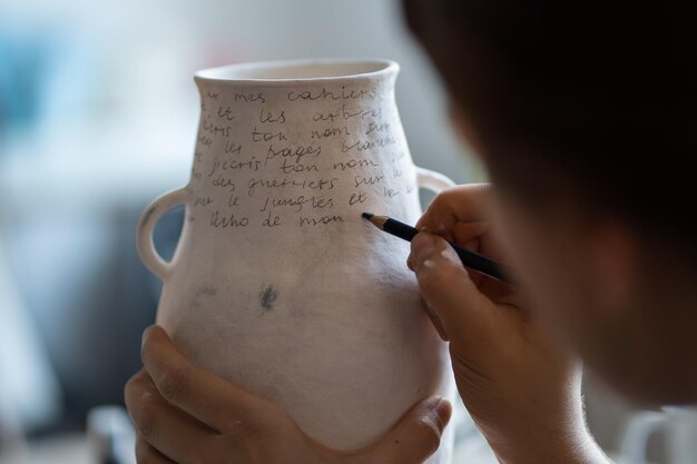 Artesã feminina pinta texto em vaso cerâmico branco com marcador em fundo desfocado na oficina