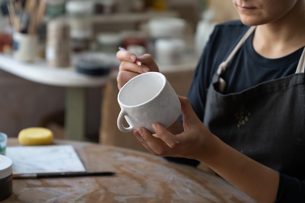 La artesana hace un boceto del patrón futuro en una taza blanca para decorar la cocina con un plato de arcilla