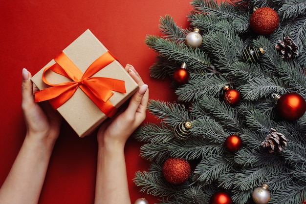 Artesanato de presente com uma fita vermelha nas mãos de uma jovem com uma manicure nua ao lado dos galhos de uma árvore de Natal.