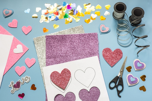 Artesanía de San Valentín, corazones y adornos de espuma y confeti sobre fondo azul.