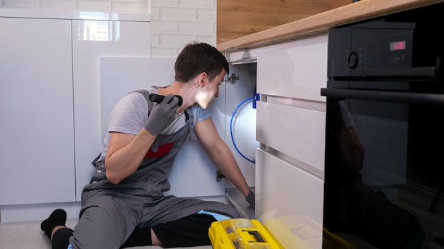 Artesano experto en mono gris y guantes con linterna incandescente examina las tuberías y la trampa de residuos debajo del fregadero en la cocina ligera contemporánea
