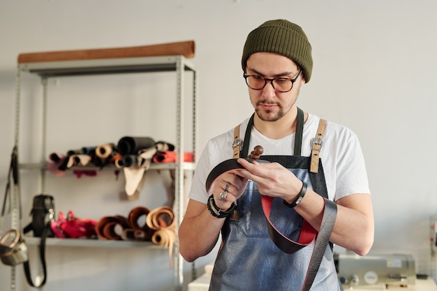 Artesano profesional en ropa de trabajo sosteniendo cinturón de cuero rojo y herramienta de mano de madera mientras trabaja sobre un nuevo artículo para el cliente