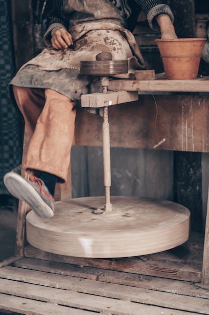 Foto artesano con ropa de trabajo sucia sentarse en la máquina de alfarero y girar la rueda con la pierna hacer vajilla en el taller de cerámica