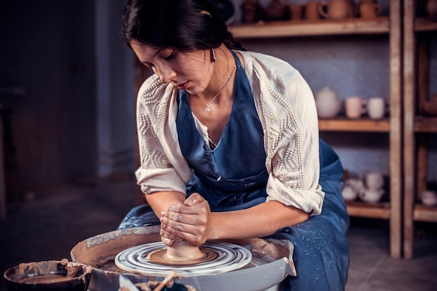 Foto artesão de oleiro elegante em sua roda criando uma nova obra-prima. ela está usando avental enquanto está sentada na cadeira.