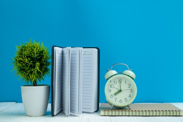 Los artículos de oficina o los artículos esenciales de las herramientas del trabajo de oficina en el escritorio de madera en lugar de trabajo, cuaderno, despertador y poco árbol con el fondo azul de la pared.