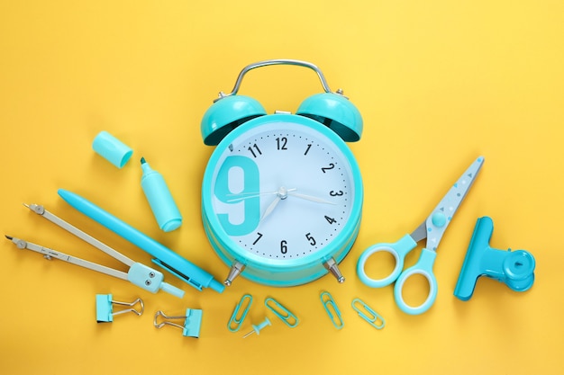 Artículos de papelería azul y reloj despertador sobre fondo amarillo. útiles escolares, tiempo para ir a la escuela, despertar por la mañana. flatlay colorido, vista superior. rotulador, bolígrafo, sujetapapeles, tijeras.