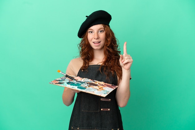 Artista adolescente ruiva segurando uma paleta isolada em um fundo verde pensando em uma ideia apontando o dedo para cima