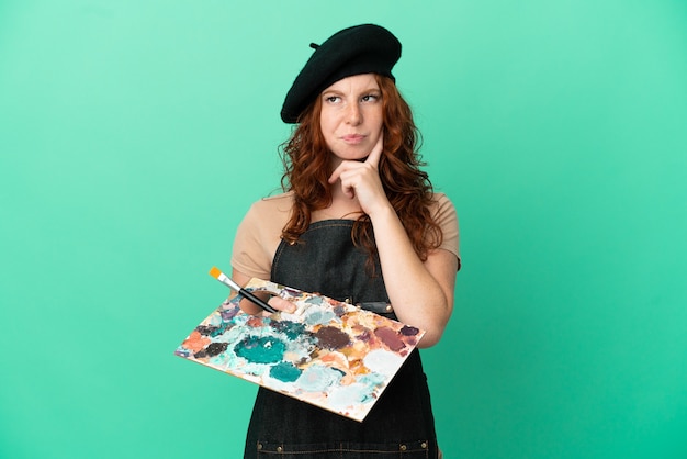 Foto artista adolescente ruiva segurando uma paleta isolada em um fundo verde, tendo dúvidas e pensando