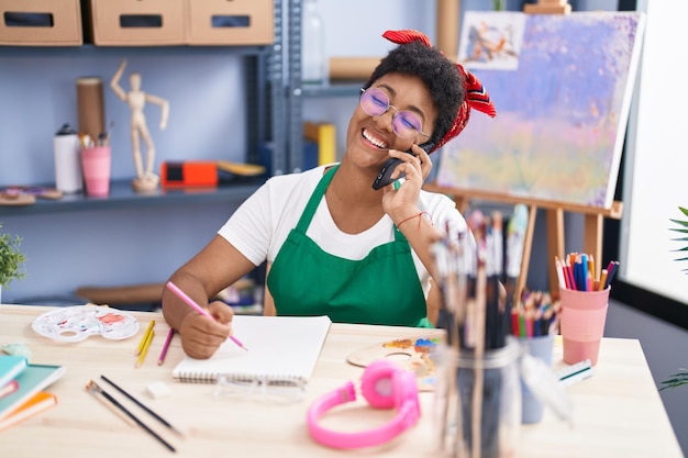 Artista afroamericana hablando en un teléfono inteligente dibujando en un cuaderno en el estudio de arte