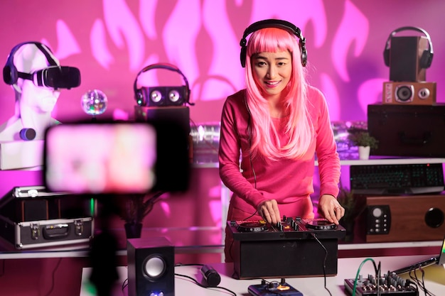 Artista alegre em pé na mesa de dj em boate tocando música techno usando console de mixagem enquanto filma sessão de música com câmera profissional. Mulher postando vídeo de performance em seu canal