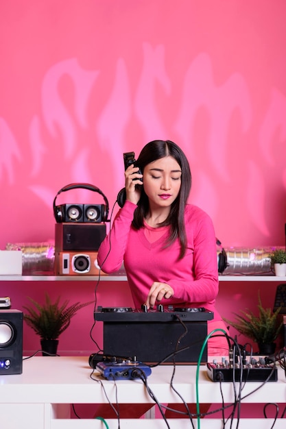 Artista asiático com fones de ouvido se divertindo enquanto toca música techno usando console de mixagem profissional, de pé na mesa de dj no estúdio com fundo rosa. Músico tocando música eletrônica no clube