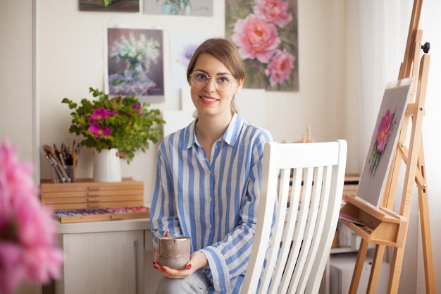 Artista caucásica hermosa niña sonriente con gafas pinta en casa studio sobre fondo de flores rosas y pinturas en la pared con artesanía para la creatividad. Concepto de creatividad e inspiración