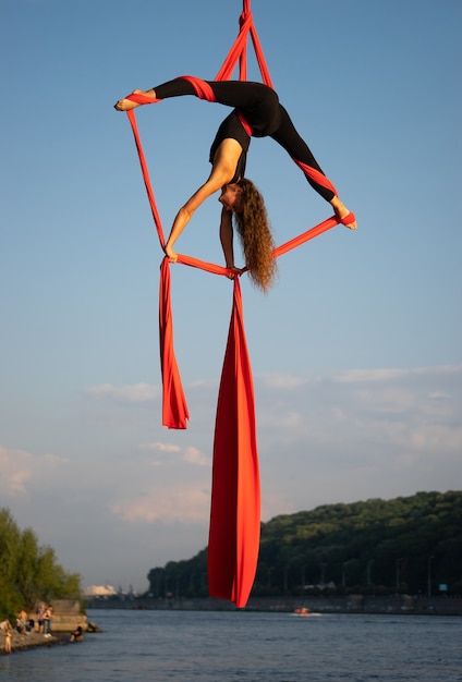 Artista de circo femenina hermosa y flexible bailando con seda aérea con el cielo y la costa del río