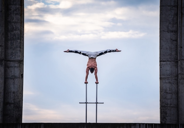 Foto artista de circo masculino flexible que hace pino contra el celaje asombroso y las construcciones modernas. concepto de simetría, equilibrio y creatividad.